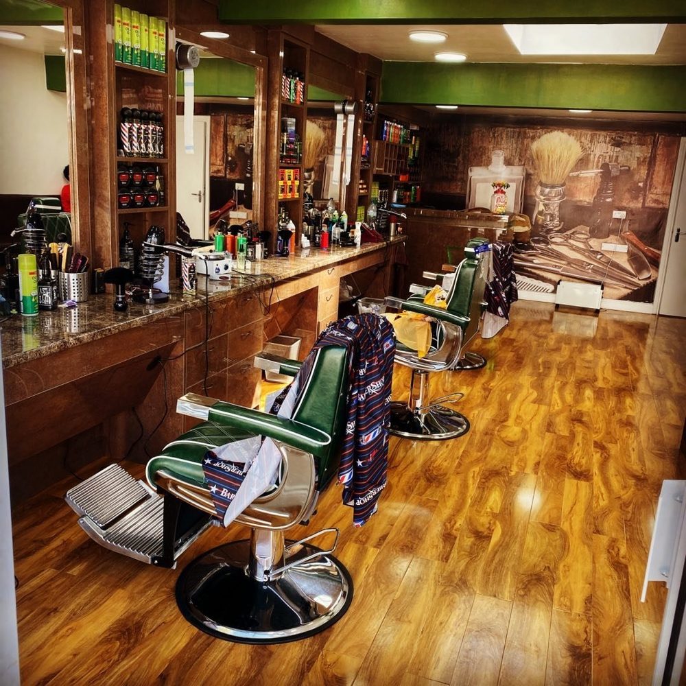 Inside the shop on High Road: two green leather barber chairs on a wood floor, with mirrors, a marble counter and shelves of products down one side.